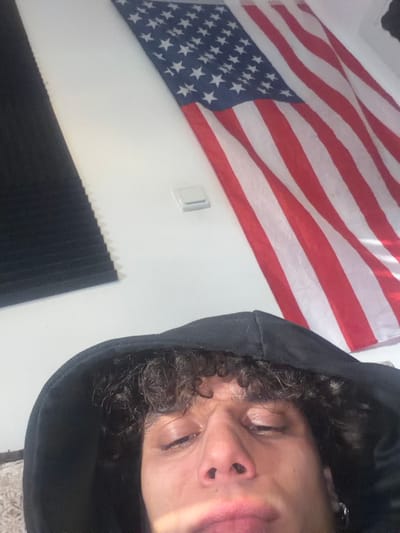 The image depicts an individual, likely male, in a room located in Migdal HaEmeq, Israel. The subject, visible from the forehead to the chin, wears a black hooded sweatshirt and has dark, curly hair. His eyes are partially closed, and a light reflection spans his lower face. A silver hoop earring is visible in his left ear. The background features a white wall. Mounted on the wall is an American flag, oriented horizontally with the blue field of stars in the upper left section of the flag. To the left of the flag and above the subject's head, a dark, corrugated acoustic panel is affixed to the wall. A white rectangular light switch is positioned on the wall below the flag and to the right of the acoustic panel.