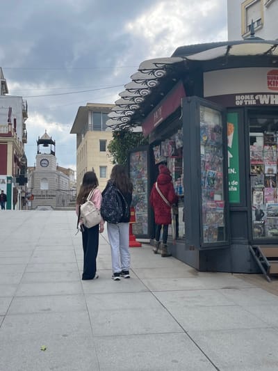 This image captures a street scene in Mérida, Spain, centered around a newsstand on a cloudy day. In the foreground, a newsstand with a distinctive striped awning, supported by ornate wrought iron, is prominently displayed. Its shelves are filled with various magazines and newspapers, some featuring titles like "HISTORIA." The side of the newsstand displays advertising text, including fragmented phrases such as "BURG KIP" (likely "BURGER KING") and "HOME OF THE WH" (likely "HOME OF THE WHOPPER"). The word "HOY" (Spanish for "Today") is also visible on its facade.
Several individuals are engaged with the newsstand. One person, wearing a red coat, is actively browsing the displayed items. Nearby, two other younger individuals with long hair are also looking towards the stand; one wears a floral patterned backpack, and the other a light-colored backpack. A fourth person is partially visible further down the street.
The street is paved with light-colored tiles, indicating a pedestrian area. In the background, an urban landscape unfolds with various buildings. A notable architectural feature is a clock tower with a dome, part of a larger building that bears text identifying it as "OFICINA DE TURISMO" (Tourist Office) and "Ayuntamiento de Mérida" (City Hall of Mérida), confirming the location. The sky is overcast, suggesting a daytime scene without direct sunlight. Overhead wires stretch across the street.
