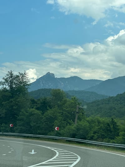 The image depicts a scenic view of a winding road in a mountainous, forested area, likely near Krivenkovskoye, Russia. The road, with white lane markings and a directional arrow, curves into the foreground. A metal guardrail separates the road from the adjacent landscape. On the left side of the road, two red and white directional signs indicate a turn. Beyond the road, a dense forest covers rolling hills, leading up to a prominent, jagged mountain peak in the distance, partially obscured by clouds. The sky is a pale blue, with scattered white clouds. A utility pole with wires is visible on the right side of the road, amidst the trees. The scene suggests a travel or roadside perspective.