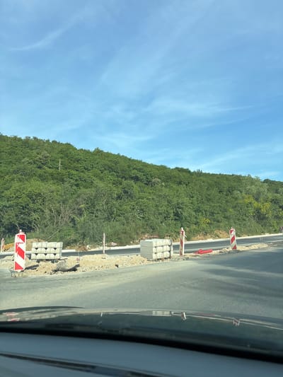 The image depicts a roadside scene in Prijeradi, Montenegro. A new asphalt road is being constructed alongside a forested hill. The roadside is marked with red and white striped barriers and stacks of concrete blocks. The sky is blue with wispy clouds. The perspective suggests the photo was taken from inside a vehicle.