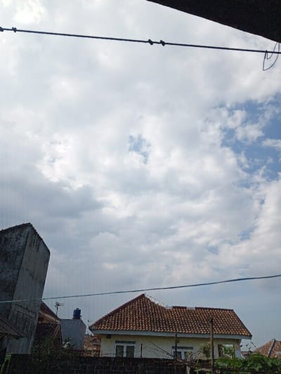 The image captures an exterior urban landscape in Weintiapa, Liberia, under a partially cloudy sky. The upper two-thirds of the frame are occupied by a sky largely covered with white and grey cumulus clouds, with discernible patches of clear blue sky in the upper right and near the center. A taut, dark wire or cable with multiple small, dark cylindrical insulators stretches horizontally across the upper part of the sky. An indistinct, dark structural element is visible in the extreme top right corner.

Below the sky, a residential area is depicted, viewed from an elevated perspective. On the left, a weathered, tall, grey concrete structure, possibly a wall or building corner, is partially visible. In the center-right, a single-story house is prominent, featuring a distinctive gabled roof covered with reddish-brown, segmented terracotta tiles. The visible portion of this house's facade is light yellow or beige, with at least two white-framed windows below the roofline. A dark, low wall or fence, topped with what appears to be a security measure like barbed wire or spikes, runs horizontally in front of the main house. Behind and to the left of this house, another building with a flat, grey roof supports a blue cylindrical water tank and a small antenna structure. A partial view of another red-tiled roof is visible to the far right. Minimal green foliage is present in the lower right foreground. No individuals, animals, or active movements are observed within the scene.