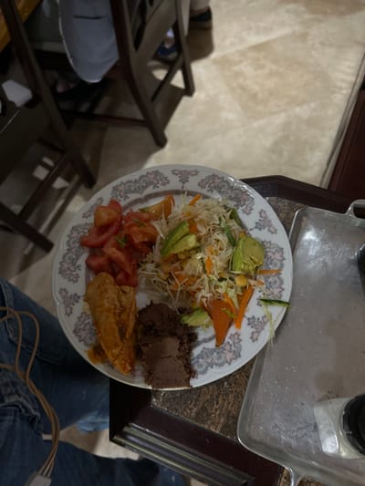 A plate of food sits on a brown wooden surface. The plate contains a salad with shredded cabbage, corn kernels, carrots, avocado, and cucumber, a portion of brown beans, and a piece of fried chicken. The plate has a floral pattern. The food is located in Río Oro, Costa Rica, at the coordinates provided. A pair of blue jeans and a brown wooden chair are visible in the foreground. The scene is dimly lit. The food appears to be a typical Costa Rican meal.