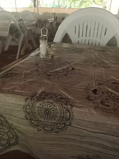 A close-up, eye-level shot shows a plastic tablecloth with a wood-grain pattern and black mandala designs. On the tablecloth, a clear glass salt shaker with a metal lid and handle sits next to a small metal object. In the blurred background, rows of white plastic chairs are visible under a covered patio area. The scene appears to be an outdoor seating area of a restaurant or cafe in Khudat, Azerbaijan.