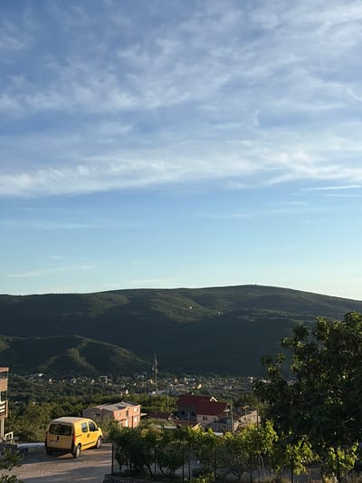 The image captures a panoramic view overlooking a mountainous landscape, likely Pelinovo, Montenegro, from a elevated vantage point on a clear day. The sky, occupying the upper left half of the frame, is bright blue with scattered, thin white wispy clouds.
The horizon line, slightly below the center, reveals a vast expanse of rolling mountains and hills, densely covered with dark green vegetation, indicating thick forests. The mountains recede into the distance, with some appearing lighter in shade due to atmospheric haze.
Nestled among the foothills and within the valleys, a town or village is visible. Various buildings with different roof colors, including prominent red-tiled roofs, are scattered across the landscape. Construction seems to be ongoing for some structures. Tall trees with lush green foliage frame the right side and foreground of the image, partially obscuring parts of the town.
In the lower right foreground, a bright yellow utility van, possibly a Renault Kangoo, is parked on a paved road or driveway. The van faces away from the viewer, angled slightly to the left. The license plate has some visible characters, but they are indistinct.
The scene is bathed in bright daylight, suggesting a sunny day, possibly late morning or early afternoon, with shadows indicating the sun is relatively high. No people are visible, and there is no direct activity taking place beyond the stationary van and the serene landscape.