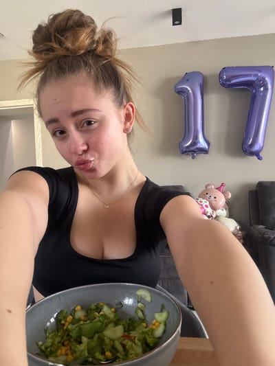 A female individual, appearing to be a young adult with light-toned skin and brown hair styled in a messy bun, poses for a selfie in an indoor residential setting in Kipra, Bulgaria. She wears a black, low-cut, short-sleeved top and a delicate gold-colored necklace with a small circular pendant. Her lips are pursed, and she looks directly at the camera; small stud earrings are visible in her earlobes. In the immediate foreground, a grey-blue ceramic bowl containing a salad of green lettuce, sliced cucumbers, and corn kernels is positioned on a light-colored wooden surface, with a metal spoon inside. The background features a light-colored wall from which two large, metallic purple foil balloons, shaped as the numbers "1" and "7," are affixed. To the right, behind the individual, various decorative items are visible on a dark upholstered surface, including a pink inflatable teddy bear head with a party hat and a light-colored plush toy. A black rectangular ceiling light fixture is mounted on the light-colored ceiling.