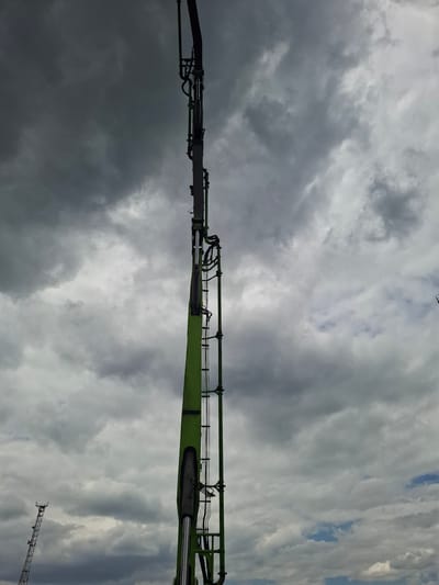 The image shows a tall, green industrial boom against a cloudy sky. The boom is partially obscured by the dark, heavy clouds which dominate the upper portion of the frame. The boom appears to be part of a construction crane or a similar piece of heavy machinery. The lower portion of the image shows a glimpse of a distant lattice tower, possibly a communication tower, also silhouetted against the sky. The overall impression is one of an overcast day in an industrial or construction setting. No people are visible in the image. The location in the city of Argun, Russia, is not visually ascertainable from the image content.