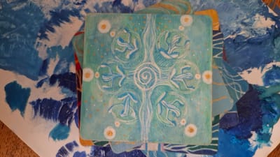 The image displays a centrally positioned, square-shaped, hand-painted panel or tile with slightly rounded corners. The panel features a light teal background adorned with a symmetrical, stylized design in white and light blue. At its core is a white spiral, from which an elongated, undulating white form extends vertically. Surrounding the central spiral are eight stylized, curvilinear leaf-like or floral motifs rendered in white and shades of blue, arranged in a circular pattern, with some enclosed within white circular outlines. The teal background is dotted with numerous small orange or gold flecks, and four larger white circles with orange centers are positioned near the cardinal points of the design.
This main panel rests upon a layered arrangement of other painted materials, primarily sheets of paper or cardstock. These underlying elements are characterized by abstract, textured brushstrokes in various shades of blue (from dark navy to light sky blue) and white, suggesting a vibrant artistic palette or a collection of painted scraps. Some pieces show hints of gold or yellow lines. A small section of a light brown, possibly wooden, surface is visible at the bottom-left corner of the frame. There are no people, animals, or direct actions depicted. The scene contains no identifiable geographical markers to indicate a location in Rassvet, Russia.