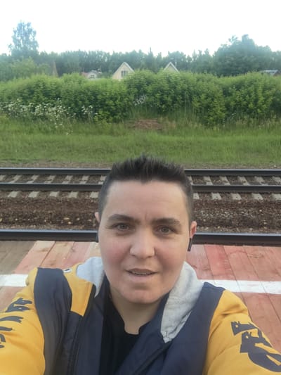 A medium shot captures a person from the chest up, standing on a wooden platform next to train tracks. The person has short, dark hair and is looking at the camera with a neutral expression. They are wearing a blue and yellow jacket over a black shirt and a grey hood. Two black earbuds are visible, one in each ear. In the background, there are lush green bushes and trees, with glimpses of two houses peeking through the foliage. The train tracks stretch across the middle ground, and beyond them, a grassy area leads to the dense greenery. The scene appears to be outdoors in a semi-rural or suburban setting, possibly a train station in Цибино, Russia.
