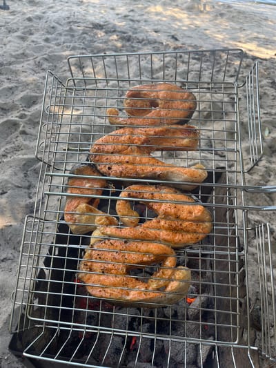 In Demydiv, Ukraine, on a sandy embankment, likely near a body of water given the presence of sand, several salmon steaks are grilling on a metal barbecue basket over hot coals. The steaks are arranged in two layers within the basket. The background shows more sand and hints of water. The scene is captured from a slightly overhead perspective.
