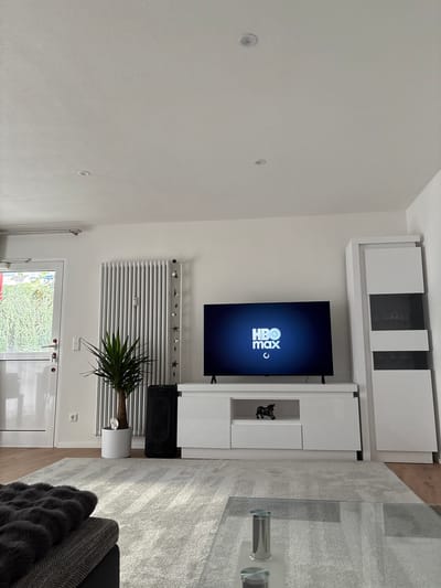 This media file captures a modern living room in Forbach, Germany. The scene is set indoors, with a prominent wall-mounted entertainment unit and a large television displaying the HBO Max logo. The room features clean white walls, with recessed ceiling lights. A white radiator is mounted on the wall, adorned with a string of decorative stars. To the right of the radiator, a green potted plant with long, slender leaves adds a touch of nature to the space. A black speaker is placed on the floor to the left of the plant.
Adjacent to the entertainment unit is a glass-top coffee table with a textured grey rug underneath. A dark grey sofa with a fluffy throw blanket is partially visible on the right side of the image. The overall impression is one of a well-lit, contemporary living space. The image appears to have been taken during daylight hours, though there are no direct cues about the time of day or weather conditions outside. The location is identified as Forbach, Germany. The primary objects in focus are the entertainment system, television, and surrounding furniture, arranged in a tidy and organized manner. No people are visible in the frame.