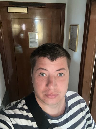 A man, appearing to be in his late 30s or early 40s, takes a selfie in what appears to be a hotel hallway. He has short, dark hair, light skin, and green eyes. He is wearing a navy and white striped t-shirt. The background shows a dark brown wooden door with a brass door frame and a small framed notice on it. To the right, a light-colored wall is visible with another framed notice. The flooring is not visible. The scene is lit by ambient lighting, likely from within the hotel. There is no explicit indication of the specific city, San Martino Buon Albergo, Italy.