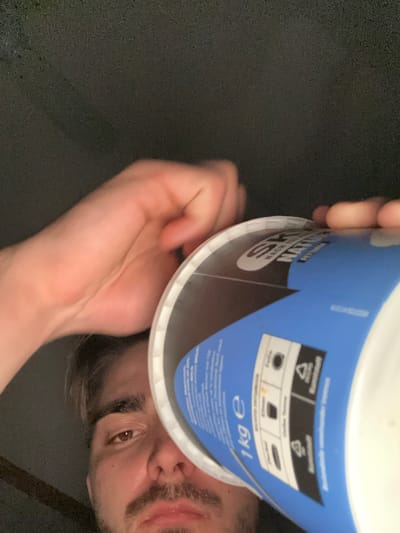A man holds a blue and white container in front of his face. The container appears to be a 1 kg product, with recycling symbols visible on the side. The man has brown eyes, dark hair, and a mustache. He is holding the container with both hands. The background is dark, and it is unclear if the location is in Rafz, Switzerland.