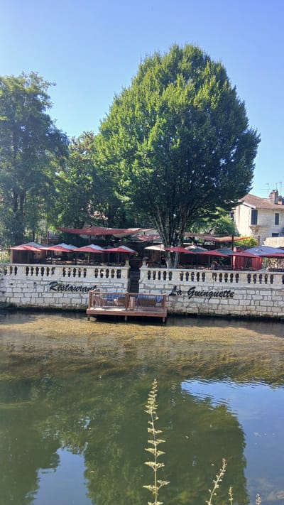 The image shows the outdoor seating area of a restaurant called "La Guinguette" located in Brantôme en Périgord, France. The restaurant is situated along a canal or river, with clear water reflecting the surrounding trees and sky. The restaurant features a stone wall with decorative railing, behind which are several tables and chairs covered by red and white umbrellas. A wooden deck with seating is in the foreground, extending over the water. Tall trees with lush green foliage are visible behind the restaurant, and a building with a tiled roof is visible in the background. The sky is clear and blue. No people are clearly visible in the scene.