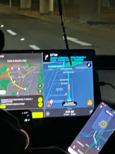 The image shows the navigation system on two electronic devices, possibly in a car. One device appears to be a tablet or large phone displaying the navigation route in Pudahuel, Chile, while the second is a smartphone showing a similar map. The route shows a travel time of 21 minutes over a distance of 25 km. The destination is Aeropuerto Arturo Merino Benítez, located at 11 National. The suggested route involves Autopista Américo Vespucio and Avenida Armando Cortínez. The phrase "Ponte el cinturón y listo" ("Put on your seatbelt and you're ready") suggests the driver or passenger is about to start the journey. Also visible is an Apple Watch.