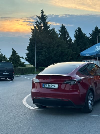 The image depicts a parking area in Momkovo, Bulgaria, under a sky exhibiting sunset or sunrise colors. In the foreground, a red Tesla Model Y is parked, angled slightly towards the right. Its rear is visible, showing a dark-tinted rear window, a subtle integrated spoiler, and a prominent "T" logo. The vehicle carries a Bulgarian license plate "EA 8899 AB". The right rear wheel with multi-spoke alloy rims is visible. Further back on the left, a black Volkswagen van is parked, displaying its rear with a tow hitch and Bulgarian license plate "K 2813 BK". Both vehicles are situated on an asphalt surface, which features white painted parking lines, including a curved line around the rear of the Tesla and a straight line near the Volkswagen van. The background consists of a dense line of tall evergreen trees, fronted by a low green hedge. A black street light pole stands between the Tesla and the trees. On the far right, a blue umbrella is partially visible, displaying text that includes "DEVIN Mineralna & Vitaminna", suggesting a nearby kiosk or cafe. The sky transitions from a distinct band of orange and yellow clouds near the horizon on the left to lighter blue and grey clouds overhead. No individuals are present in the scene.
