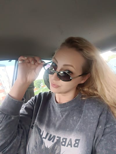 A blonde woman is captured in a close-up, selfie-style photograph, seated inside a car in Тоншалово, Russia. She looks directly towards the camera, a slight smirk playing on her lips. Her light-colored hair flows loosely around her shoulders and back. She wears dark, oval-shaped sunglasses with thin metallic frames, holding them slightly down with her right hand to partially reveal her eyes. A simple ring adorns her right ring finger.

She is dressed in a casual dark grey, long-sleeved sweatshirt that features a faded or distressed texture. Prominently displayed across the front of her top is mirrored text, which appears to spell "BALENCIAGA" in reverse.

The interior of the car is visible, with the beige ceiling and parts of the window frames and seat structure forming the background. Through the car's windows, a daytime scene is observed. On the left, a light-colored, multi-story residential building with numerous windows stands against a bright, possibly overcast, sky. Green foliage and trees are also visible outside the windows, suggesting a suburban or urban setting. The overall impression is a casual, relaxed moment.
