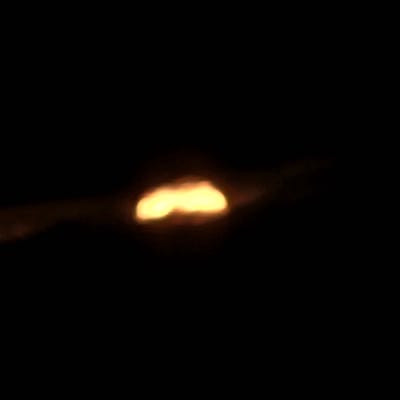 The image displays a highly blurred, elongated, irregular orange-yellow light source positioned horizontally against a black background. Subtle, darker reddish-brown streaks or diffused light extend horizontally from the central illumination. No discernible individuals, specific objects, or detailed environmental features are visible due to extreme darkness and lack of focus. The scene is located in Nzalat Laadam, Morocco.