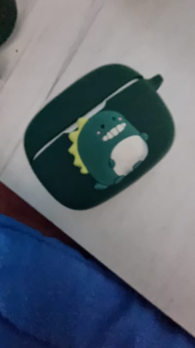 A close-up, slightly out-of-focus, and slightly angled shot shows a dark green case with a cartoon dinosaur figure on a light surface. The case appears to be for an electronic device, possibly earbuds, featuring a small loop at the top. The cartoon dinosaur is primarily dark green with a lighter, cream-colored belly and a yellow frill. Part of a blue item (possibly clothing or a blanket) is visible in the lower right corner. The location is undetermined.