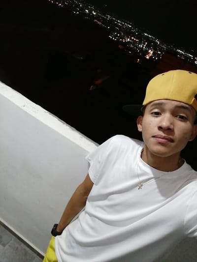 A young man wearing a yellow baseball cap, a white t-shirt, and a gold necklace stands in front of a white wall or railing. A watch is visible on his left wrist. Behind him is a dark expanse with scattered lights, possibly a city at night seen from a height. The lighting is dim, with the man illuminated from the front.