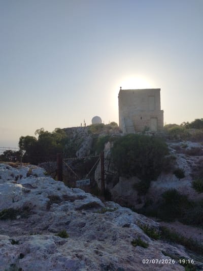 The image depicts a rocky, arid landscape at sunset, with a sun-drenched, stone tower as the central object. The tower, appearing to be a historic structure, is built on a rocky outcrop and has two visible doorways and an external staircase. In the background, a large white radar dome is visible on the horizon, along with a few indistinct figures standing near it. The sun is positioned behind the tower, creating a bright lens flare effect and casting a warm glow across the sky, suggesting it is either sunrise or sunset. The vegetation is sparse, consisting of scrubby bushes and some trees. A metal gate with a "PRIVATE LAND NO ENTRY" sign is visible in the foreground, indicating restricted access to the area. The overall mood is serene and perhaps a little remote. The timestamp at the bottom right corner indicates the date as 02/07/2026 and the time as 19:16, confirming the evening setting. The location context suggests this is likely Dingli, Malta, a place known for its dramatic cliffs and historical sites.