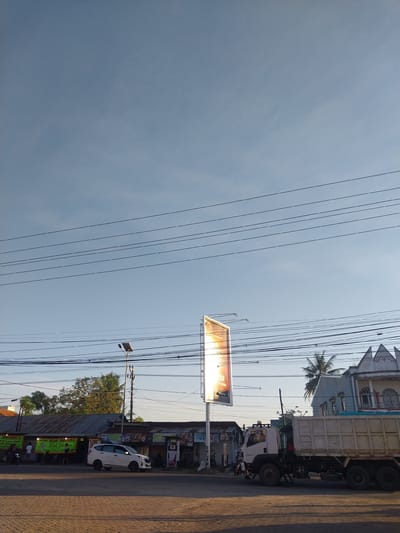 The media file displays a street scene in Macege, Indonesia, bathed in the warm glow of late afternoon sun. The image is oriented vertically, with power lines crisscrossing the sky, creating a sense of urban infrastructure against the clear, pale blue sky.

Dominating the right side of the frame is a large, white dump truck, parked on the side of a paved road. In front of the truck, a white car is also parked. A billboard with a brightly lit advertisement stands prominently near the road, its illumination suggesting it is either evening or the advertisement is internally lit. Further down the road, a low-rise building with a corrugated metal roof appears to be a shop or eatery, with several motorcycles parked in front. Signs are visible, indicating business activities. One sign, in yellow and green, reads "NASI GORENG" (fried rice) in Indonesian. Another sign, partially visible, reads "MAX JUARA" (Max Champion).

The presence of palm trees and the style of architecture, particularly the building behind the dump truck, hint at a tropical, Southeast Asian setting. The sunlight casts long shadows, reinforcing the indication of late afternoon. There are no visible people in focus, but the parked vehicles and the commercial signs suggest a lively, inhabited area. The overall mood is one of everyday activity in a bustling street.
