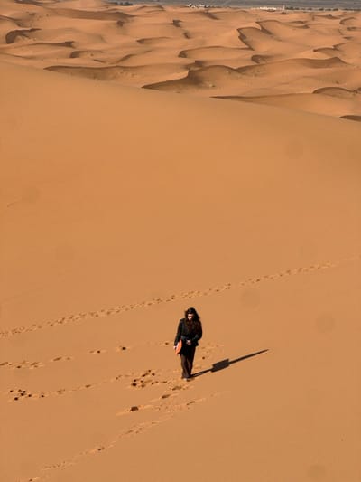 A solitary individual walks across a vast expanse of reddish-orange sand dunes in the Erg Chebbi desert, near Rissani, Morocco. The person, appearing female with long dark hair, is dressed in a dark jacket and trousers, and holds a bright orange, disc-shaped object in her left hand at waist level. She is oriented slightly to her left, looking downwards, and casts a distinct, elongated shadow towards the lower right, indicating illumination from the upper left. A trail of footprints extends across the sand, originating from behind her and continuing along her path. In the far background, a faint horizon reveals distant low-lying structures and sparse vegetation, indicating the proximity of a settled area.