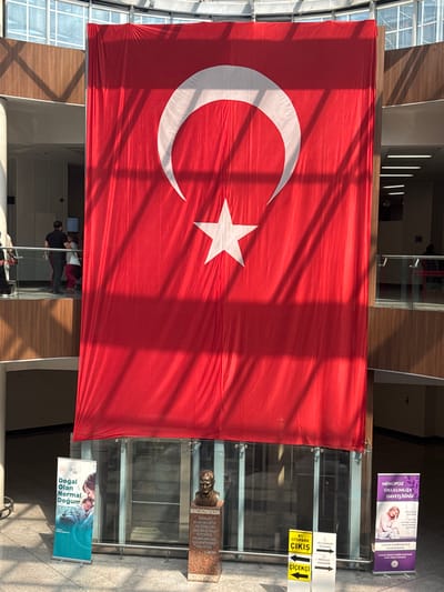 A large Turkish flag is displayed prominently, hanging from a ceiling and reaching down towards the ground. The flag features a white crescent moon and a white star on a red background. Below the flag, a bust of Mustafa Kemal Atatürk is mounted on a pedestal. To the left of the bust, a vertical banner with the text "DOĞAL OLAN NORMAL DOĞUM" (Natural Birth is Normal Birth) is displayed. To the right of the bust, another vertical banner reads "MENOPOZ OKULUMUZA DAVETLİSİNİZ" (You are invited to our Menopause School). Multiple directional signs are visible, including "B1 OTOPARK" (B1 Parking), "ÇIKIŞ" (Exit), and "ÇİÇEKÇİ" (Florist). In the background, people are seen walking on an upper level, and the environment appears to be an indoor public space with architectural elements such as wooden panels and large windows. The presence of the Turkish flag and the bust of Atatürk suggest a patriotic or memorial context, possibly within a public building in Çankaya, Turkey.