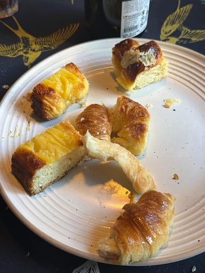 The image shows a plate of pastries on a table. The pastries include croissant-like items and square slices of glazed bread. The plate is white with a speckled pattern and raised concentric rings. The table is covered with a black tablecloth decorated with gold bird patterns. A bottle is partially visible on the left side of the plate. The location is La Lucila, Argentina.