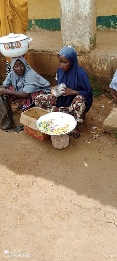 In Kagarko North, Nigeria, two young women are seated on the ground, engaged in what appears to be a transaction. One woman, on the left, wears a light blue headscarf and a grey top, with a large white pot balanced on her head. She is looking to her right with a concerned expression. Beside her is a black bag. The second woman, on the right, wears a dark blue headscarf and a patterned skirt. She is holding a clear plastic container filled with small, individually wrapped candies. In front of them is a white tray containing more of these candies, spread out for display. A cardboard box, likely used for packaging, is situated behind the tray. The ground beneath them is a mixture of dirt and scattered debris. The background consists of a weathered wall with a green and yellow stripe, and a concrete step. The lighting suggests it is daytime.