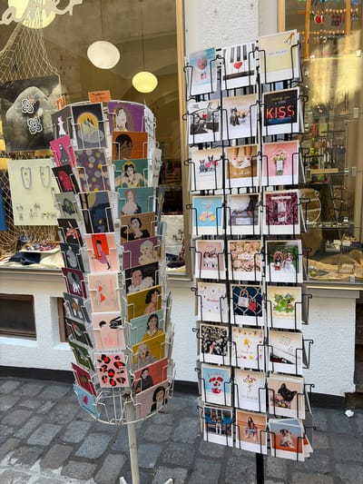 A postcard display rack stands on a cobblestone street in front of a shop window in Würzburg, Germany. The rack is filled with various postcards, many featuring artistic illustrations or photographs. Some postcards have German text on them, such as "Glückwunsch" and "Danke." Behind the rack, the shop window displays jewelry, ornaments, and a net. The exterior of the building is white stucco. The image is taken from a slightly elevated perspective.