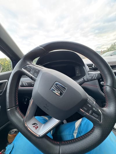 The image captures the interior of a SEAT FR vehicle from the driver's perspective, located in Kaysersberg Vignoble, France. The foreground is dominated by a black leather steering wheel with red contrast stitching, displaying a central 'SEAT' emblem and an "FR" badge on the lower spoke. Multi-function controls are present on both sides of the steering wheel. Behind it, a digital dashboard and air vents are partially visible. A wiper/washer stalk extends from the right of the steering column. The driver's lap, clad in bright blue trousers, is visible at the bottom of the frame, with a partially consumed amber-colored beverage can resting near the left thigh. The exterior environment, viewed through the windshield, shows an overcast sky, green foliage to the left, and a partially visible vehicle with distant structures or hills in the background.