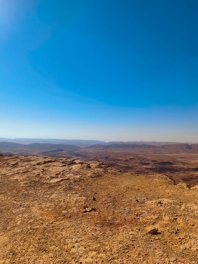 The image displays a vast desert landscape under a clear, bright blue sky. In the foreground, the terrain is rocky and arid, with shades of orange and brown dominating the ground. The rocks appear weathered and uneven. Beyond the immediate foreground, the land slopes down into a wide valley or crater, characterized by a textured, sandy, and rocky surface. In the distance, a range of rolling hills or mountains forms the horizon, appearing muted and hazy under the daylight. The scene is devoid of any human presence or man-made structures, conveying a sense of natural, undeveloped wilderness. This geological formation is likely the Ramon Crater, a prominent natural landmark located in the Negev Desert, near the city of Yeruham, Israel.