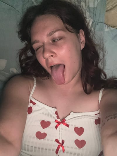The image shows a close-up, overhead view of a person lying down, with their head turned towards the camera. The person has dark hair and is wearing a white top adorned with red hearts and red bows. Their tongue is sticking out. The background appears to be a bed with a teal-colored blanket. The lighting is dim, suggesting it may be nighttime or an indoor setting with low light. No text is visible in the image. The overall mood is playful or provocative. The location context is Bishkek, Kyrgyzstan.