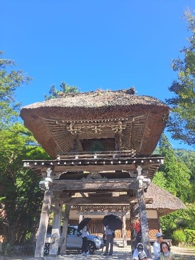This image displays a multi-tiered traditional Japanese *shōrōmon* (bell tower gate) featuring a thick, multi-layered thatched roof, located in Shirakawa, Japan. The structure is constructed from dark, weathered wood with exposed joinery. The upper tier contains a dark, bell-like object. A clear, bright blue sky is visible above the gate. Lush green trees and dense foliage surround the gate, with forested hills present in the background. Beneath the gate's archway, a light-colored SUV is parked. Six individuals are visible: three on the left, two of whom hold large black umbrellas for sun protection; and three on the right, one partially obscured by a pillar, another seated, and a third wearing sunglasses. The ground beneath the gate appears to be unpaved compacted earth or gravel.