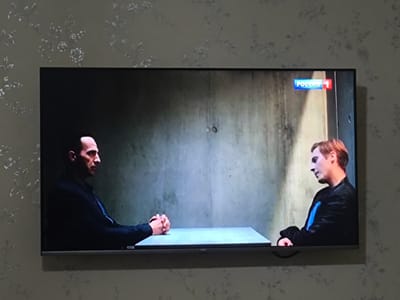 The image shows a television screen displaying a scene with two men seated at a table in front of a concrete wall. The man on the left, facing right, is wearing a dark jacket and has his hands clasped in front of him on the table. The man on the right, facing left, is wearing a black jacket with a blue stripe and appears to be looking downwards. A logo for "Россия 1" is visible in the top right corner of the television screen. The television is mounted on a wall with floral wallpaper. The scene is consistent with a filmed interview or interrogation.