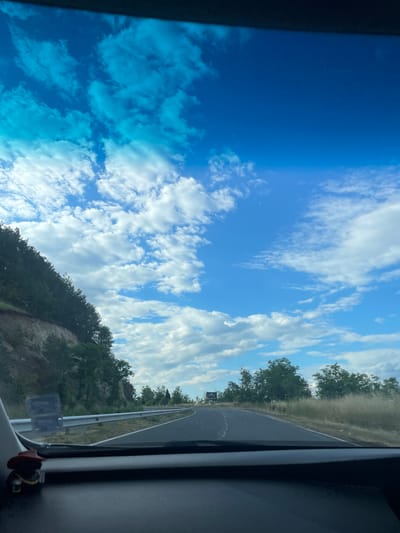 The image depicts a view from inside a vehicle, looking forward through the windshield. The road ahead curves to the right, bordered by a grassy shoulder and a metal guardrail on the left. A steep, wooded embankment rises along the left side of the road, with dense green trees covering its slope. The sky is a vibrant blue, filled with scattered white and light blue clouds. The dashboard of the vehicle is visible in the foreground, with a small, dark figurine wearing a brown hat placed on the left side. The scene appears to be a scenic drive on a highway outside of Balabanovo, Bulgaria, on a clear day.