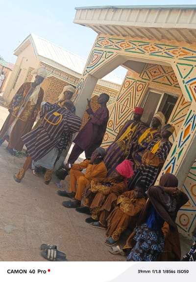 A group of people are gathered outside a building adorned with geometric patterns in Wase, Nigeria. The building has a white roof and the geometric patterns are painted in shades of brown, yellow, blue, and green.

The people are dressed in a variety of traditional clothing. Some wear robes and head coverings, while others wear long-sleeved shirts and dresses. One man is standing and pointing. Some people are sitting, while others are standing. Two discarded pairs of sandals are on the ground.
