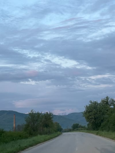 The image captures a serene landscape in what appears to be either early morning or late evening light. The sky, dominating the upper portion of the frame, is largely overcast with white and light grey clouds, streaked with very faint hints of pink or orange, suggesting a low sun angle.
Below the sky, a natural and semi-rural environment unfolds. A paved road curves gently into the distance on the right side of the image. The road is bordered by vibrant green grass and dense trees, with foliage visible in various shades of green, indicating healthy vegetation. In the background, large, dark green mountains or hills rise prominently, their upper slopes partially veiled by the soft light and atmospheric haze.
A notable man-made object is present: a tall, slender, reddish-brown cylindrical structure, resembling a brick chimney or smokestack, stands against the mountain backdrop to the left of the road. No people or specific activities are visible, contributing to a tranquil and still atmosphere. The overall scene conveys a quiet moment in nature, with subtle hints of human presence. The location is Gorna Rositsa, Bulgaria. No visible text is present.