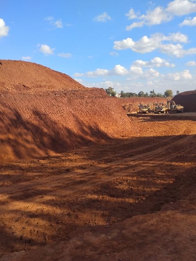 This image depicts a large-scale earthmoving and excavation site located in Kikuyu, Kenya. The predominant feature is reddish-brown soil, characteristic of lateritic earth, which forms large, uneven mounds and excavated depressions across the scene. The foreground shows disturbed ground with visible vehicle tracks and strong shadows, indicating direct sunlight from the right. In the midground, a significant trench or cut-out area is flanked by steep, high piles of loose reddish-brown soil. Within this excavated zone, a cluster of three yellow heavy construction machines, identified as excavators or loaders, are parked and appear inactive. In the distant background, above the construction machinery, a line of green trees and several light-colored buildings are discernible. The sky is blue with scattered white cumulus clouds. No people are visible within the frame.