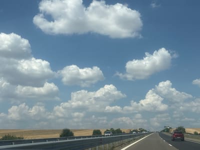 The image captures a view from a vehicle traveling on a highway under a bright blue sky with scattered white cumulus clouds. The highway has multiple lanes, with several cars visible in the distance. To the left of the highway, a dry, golden field stretches out, bordered by a line of green trees. A metallic guardrail runs along the side of the road. The overall impression is of a sunny day on a rural highway, possibly in a region with agricultural landscapes. Based on the provided metadata, this scene is located in the vicinity of Troyanovo, Bulgaria.