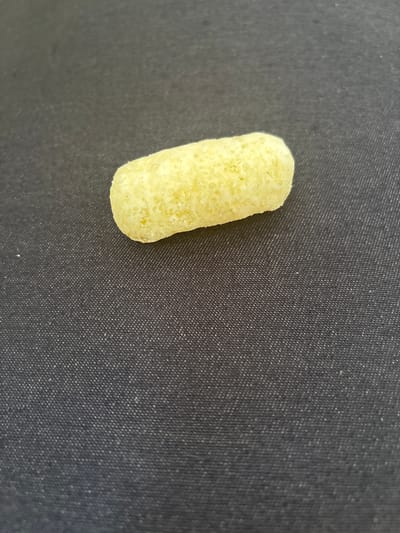 The image shows a single, pale yellow, oval-shaped puffed snack food item resting on a dark gray textured surface. The surface appears to be fabric with a visible weave pattern. The snack is centrally located in the upper half of the frame, with ample space around it. There are no people, actions, or interactions depicted. The lighting is even, with no harsh shadows. The background is out of focus, drawing attention to the snack.