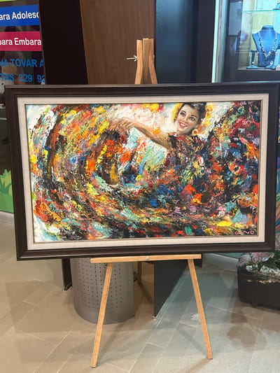 An indoor scene within a commercial or public building is depicted, likely in Plaza Vela, Boca del Río, Mexico. The primary subject is a large, rectangular oil painting displayed centrally on a light-colored wooden tripod easel.
The painting, rendered with heavy impasto, depicts an impressionistic or expressionistic smiling female figure from the upper torso, seemingly dancing or in motion. Her form and dress are composed of vibrant, swirling strokes of multi-colored paint, including reds, oranges, yellows, blues, blacks, greens, and whites. The artist's signature, "CANO," is visible in the lower right corner of the canvas. The artwork is enclosed within a dark brown frame, featuring an inner light-colored mat.
The background elements provide context for the location. To the left of the painting, a vertical sign, partially obscured, is visible. It features text in Spanish, including fragments "bara Adolesc" and "bara Embara," along with "DRA. ANA TOVAR A." and a partial phone number beginning "229 92". This sign refers to gynecological and obstetrics services, specifically mentioning adolescent pregnancy ("embarazo adolescente"), indicating a medical practice. To the right of the painting, a glass display case showcases various jewelry items, including necklaces with blue stones, suggesting the presence of a retail jewelry store. A potted plant with dark green leaves and some reddish tones is visible below the jewelry display. The floor is covered with light-colored square tiles.
The specific signage for "Dra. Ana Tovar A." with the Veracruz area code "229" and services related to adolescent pregnancy strongly suggests the location is within Plaza Vela, Boca del Río, where her practice is known to be situated. No individuals are visible in the scene.