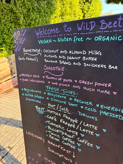 A black chalkboard sign displays a handwritten menu of food and drinks at "Wild Beet," a vegan, gluten-free, and organic establishment. The sign is positioned outdoors on a sidewalk, with dappled sunlight and shadows indicating it is daytime. The background features lush green foliage and a hint of a building. The text is written in various colored chalk, including purple, pink, green, and white.
The menu is divided into sections: "Welcome to WILD BEET," "HOMEMADE: COCONUT AND ALMOND MILKS (AND FAMOUS) ALMOND AND PEANUT BUTTER BANANA BREAD AND SNICKERS BAR," "SMOOTHIE:", and "FRESH JUICES:". Under "SMOOTHIE:", options include "PROTEIN PLUS," "PURE INDULGENCE," "FOUNTAIN OF YOUTH," "LOVE PUNCH," and "GREEN POWER." Under "FRESH JUICES:", there are categories like "* REJUVENATE," "* ULTRA GREEN," "SIGNATURE....," "* REHYDRATE," "* IMMUNE SHOT," "* RECOVER," "* COLD," "* ENERGIE," and "PRESSED."
Below this, a section labeled "BEST. HOT/COLD.... DRINKS" lists items such as "MATCHA FRAPPÉ / LATTE," "CAFÉ FRAPPÉ / LATTE," "TURMERIC LATTE," "BULLETPROOF COFFEE," "TONIC COFFEE," and "REISHI COFFEE." The sign also features decorative elements like small hearts and dots. At the bottom, partially obscured, are the words "LEGENDARY BREAFKAST," and "SCRUMPTIOUS."
The sidewalk is paved with grey square tiles. The overall impression is a vibrant, health-conscious eatery offering a variety of fresh and homemade items.
