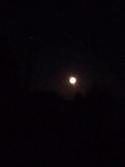 A full moon shines brightly in the dark night sky, appearing as a luminous orb against the blackness. The moon is positioned in the center-left of the frame. Below the moon, a faint, dark silhouette of trees or a treeline is visible, suggesting an outdoor environment. The rest of the image is predominantly black, with some subtle texture or noise, typical of a nighttime photograph. The location is specified as Свіслацкі сельскі Савет, Belarus, indicating a rural setting. There are no people or specific objects discernible in the image other than the moon and the implied landscape. The scene depicts a tranquil and solitary night.