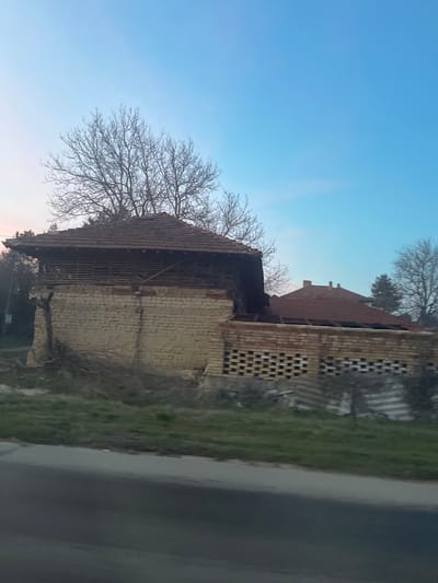 The image depicts a rural scene in Osenets, Bulgaria, featuring a dilapidated two-story structure under a clear sky. The primary building has a lower level constructed of light-colored bricks, showing signs of decay and crumbling sections. Its upper level consists of horizontal wooden slats under a gabled roof made of red clay tiles, which exhibits partial collapse and missing sections on the right side. Behind this structure, the skeletal branches of bare deciduous trees are visible. To the right, an attached or adjacent wall of red bricks features an open lattice pattern, with a partially visible tiled roof behind it, indicating another structure or extension in disrepair. The immediate foreground shows a dark asphalt road bordered by a strip of green grass and dry brush. Scattered debris, including corrugated sheets, lies within the grassy area. No individuals are present. The sky is a gradient of light blue transitioning to a faint pinkish hue near the horizon.