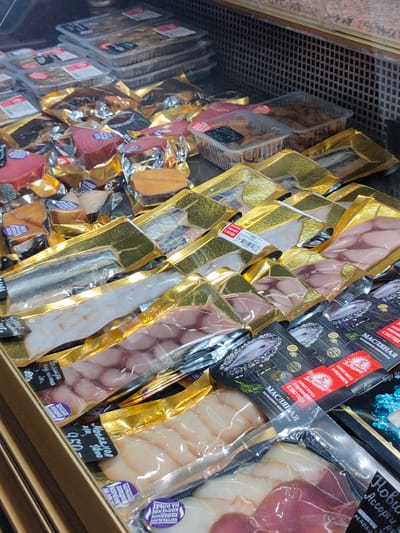 The image shows a display of packaged fish products in a retail setting, possibly a supermarket or fish market. The products are in various types of packaging, including clear plastic containers and gold foil wrappers. Some of the fish appears to be smoked or cured, with different colors and textures visible. There are price tags and labels on some of the packages, with text in Cyrillic. The background shows a grid-like structure, possibly part of a refrigerator unit or shelf. The lighting is bright, illuminating the products clearly. The location is identified as the city of Khrabrovo, Russia.