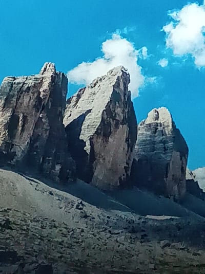 Three rocky peaks rise from a gravelly slope, their jagged forms contrasting with the bright blue sky dotted with fluffy white clouds. The leftmost peak is deeply shadowed, revealing a rugged, textured surface. The central peak is partially lit, exposing lighter gray tones and sharp edges. The rightmost peak displays layered formations and is partially shrouded in shadow. The terrain leading up to the peaks is a mix of gray and darker shades, giving the impression of a harsh, mountainous landscape.