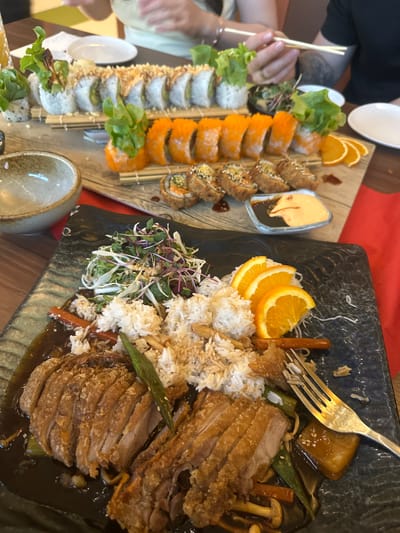 The image presents a vibrant dining scene, likely in a restaurant in Königs Wusterhausen, Germany, during the daytime. Two individuals are partially visible, with their hands suggesting they are actively engaged in eating; one hand holds chopsticks, and a wedding ring is visible on a finger.
The table features a lavish spread of Asian-inspired cuisine. On the left, a long wooden serving board displays an extensive variety of sushi rolls. These include rows of rolls topped with bright orange roe, others adorned with fresh green lettuce, and a third type appearing to be deep-fried or tempura-style, garnished with sesame seeds. Accompanying the sushi are fresh orange slices and a small dish containing two distinct sauces: one creamy orange and another dark, possibly soy-based.
On the right, a large, dark-textured plate holds a substantial main course. This dish features numerous slices of crispy-skinned meat, reminiscent of roasted duck or pork belly, served in a rich, dark sauce. It is accompanied by white rice, a fresh side salad made with mixed greens and sprouts, and an assortment of cooked vegetables such as mushrooms, green beans or okra, and carrots. More orange slices are used as a garnish on this plate, and a metal fork is visible, stuck into the rice, indicating the meal is in progress. The overall impression is of a plentiful and aesthetically pleasing meal.