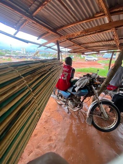 The image captures an outdoor scene, likely a temporary shelter or stall, with a person and a motorcycle as central subjects. The perspective is angled downwards, looking from above and slightly to the side.
A person, seen from the back, is seated on a light blue motorcycle. They are wearing a red jersey with the name "JORDAN" and the number "23" clearly visible on the back. The motorcycle has chrome accents and the word "DAYLONG" is visible on its engine casing. A white cloth is draped over the handlebars.
The ground beneath the motorcycle and within the shelter is a reddish-brown color, appearing somewhat wet or damp. The shelter itself has a corrugated metal roof supported by wooden beams, some of which show signs of wear. In the foreground, a large, cylindrical bundle, seemingly made of tightly woven bamboo or similar natural materials, stretches diagonally across the lower part of the frame.
Beyond the immediate shelter, the background reveals an open, earthy area with patches of green vegetation. Several cars, predominantly white and light-colored, are parked further away. In the distance, a general view of what appears to be a town or settlement is visible under a bright, overcast sky, suggesting it is daytime. A tall structure, possibly an antenna or tower, can be seen on the horizon. The overall scene suggests a functional, possibly commercial or roadside, environment in Auchi, Nigeria.