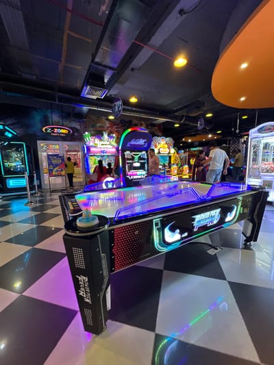The photograph captures an interior scene in what appears to be an entertainment or arcade center, possibly located in Kalyan-Dombivli, India, though this cannot be definitively confirmed from the image alone. The primary focus is a "Power Puck Fever" arcade game, standing prominently on a black and white checkered floor. The game features illuminated elements and bold lettering. Several other arcade machines are visible in the background, suggesting a bustling entertainment environment. People are seen interacting with the games in the background. Overhead, an orange ceiling structure is apparent along with industrial ductwork. The overall lighting is a mix of ambient and directed light, creating a vibrant atmosphere.